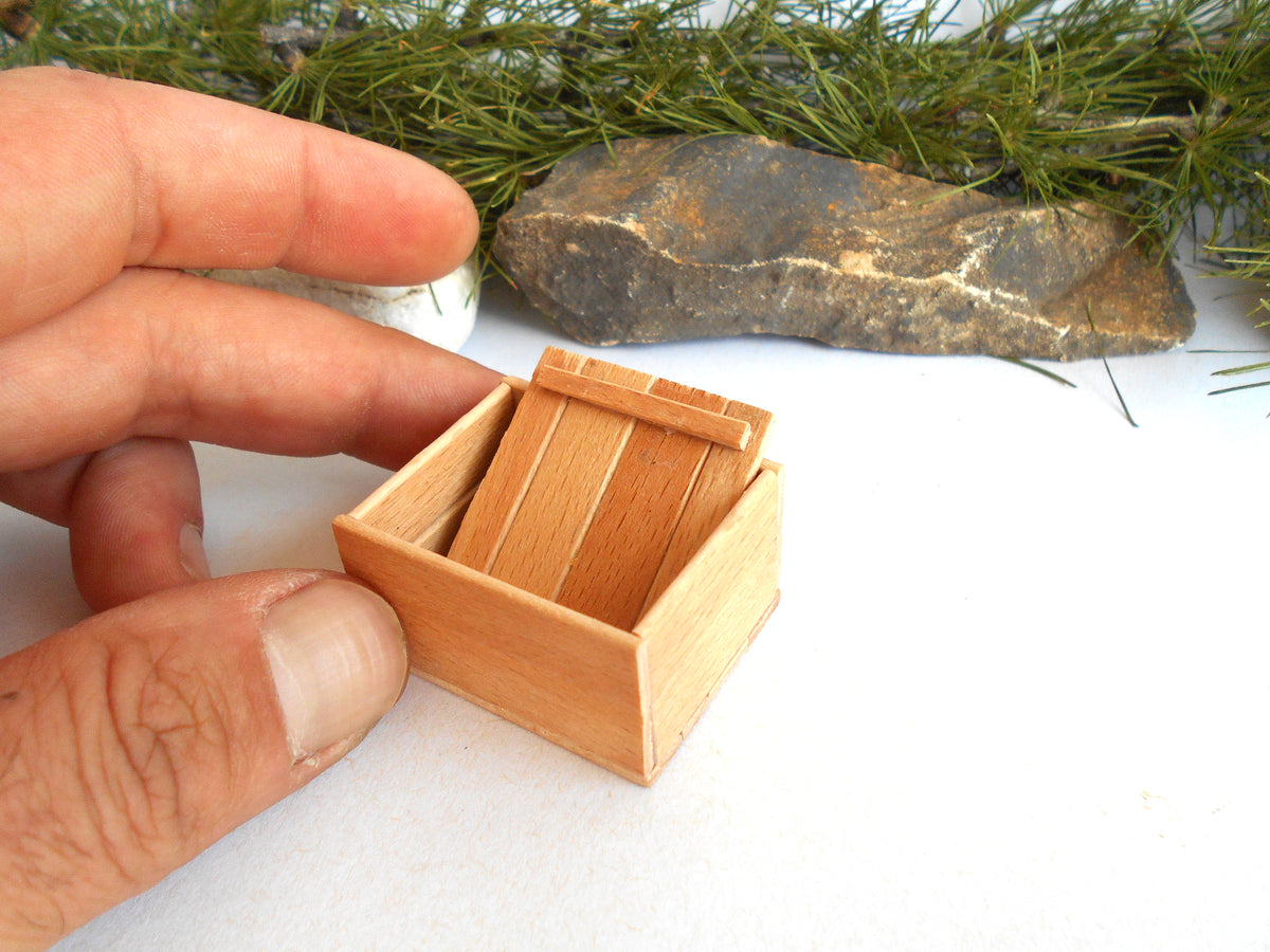 A miniature light wood box with a slatted lid, held by a person's hand against a white background with pine branches and a rock in the background.