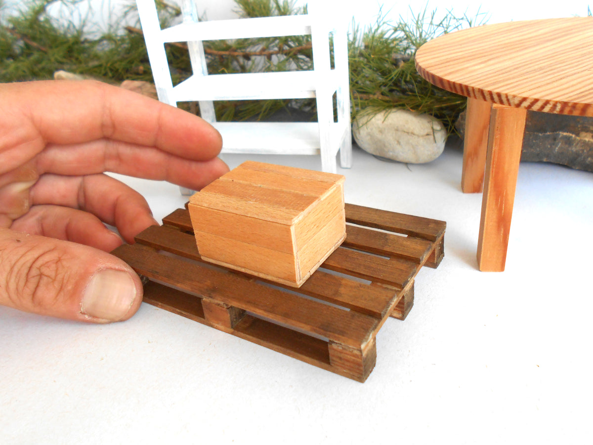 A miniature wooden box resting on a small wooden pallet, with a human hand holding it for scale in a miniature outdoor scene.