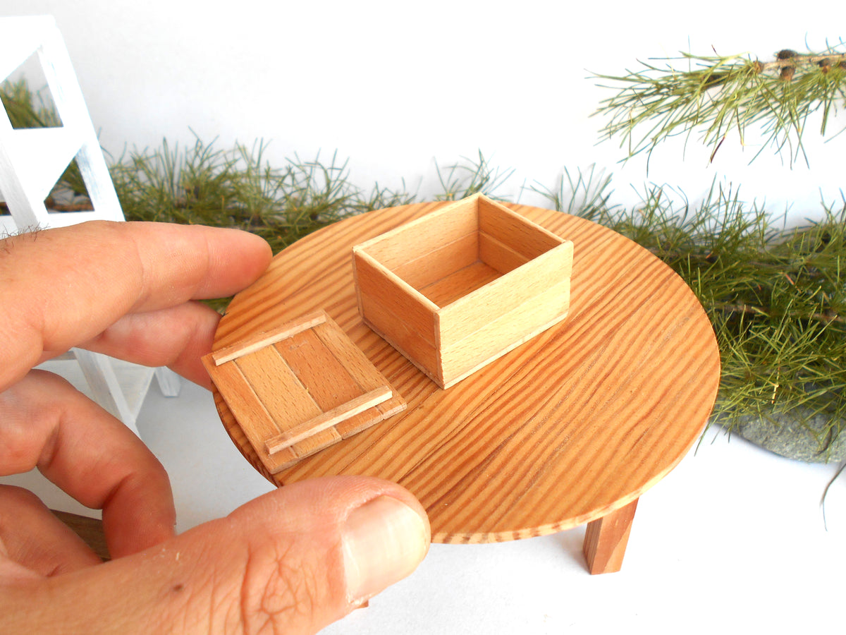 A miniature wooden dollhouse accessory set featuring a round table, a small box, and a lid, held by a hand to show scale against a white background with green foliage.