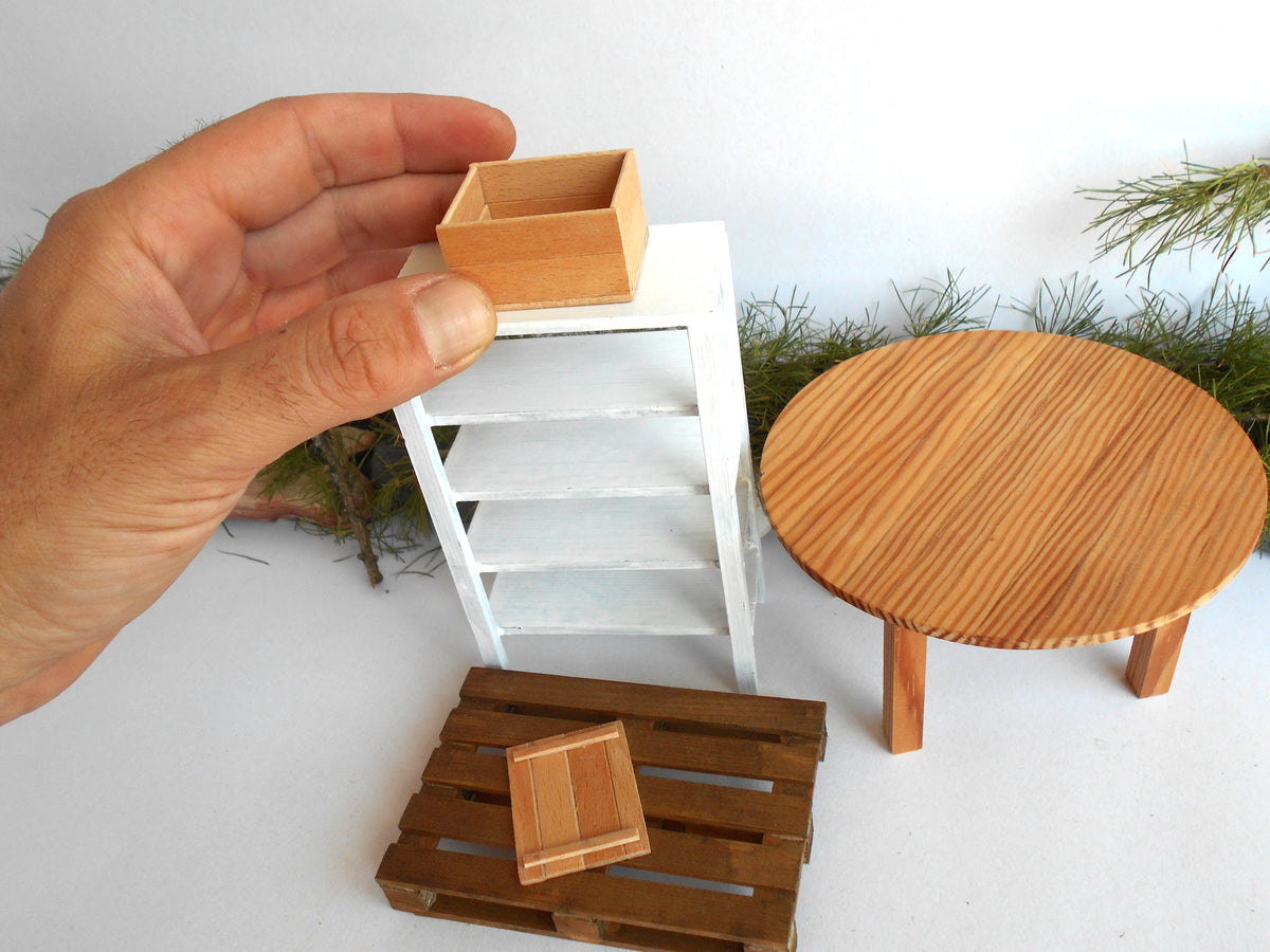 A hand holds a small wooden box above a miniature white shelf, with other tiny furniture pieces including a round wooden table and a slatted wooden crate on a white surface.