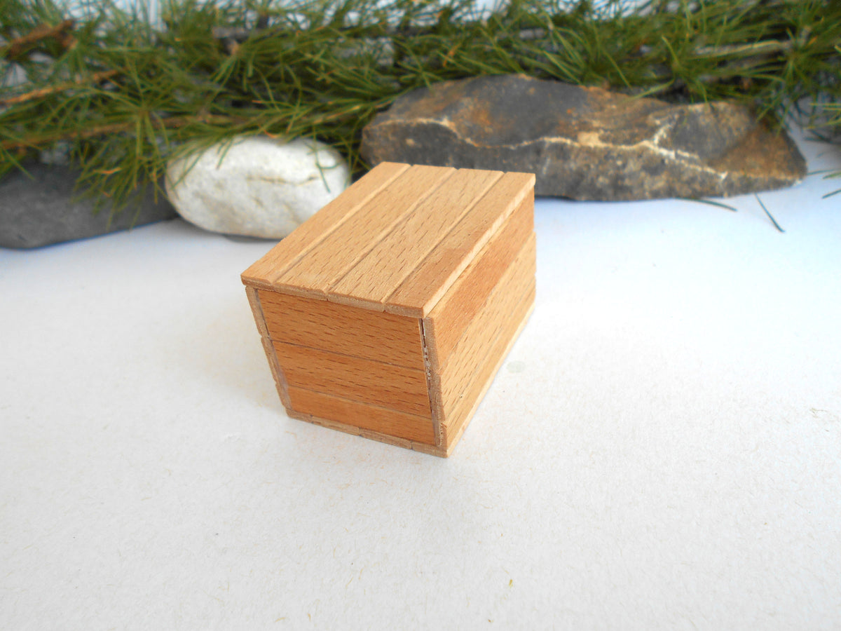 A small, handcrafted wooden box made of light-colored wood planks, set against a white surface with decorative green pine branches and rocks in the background.