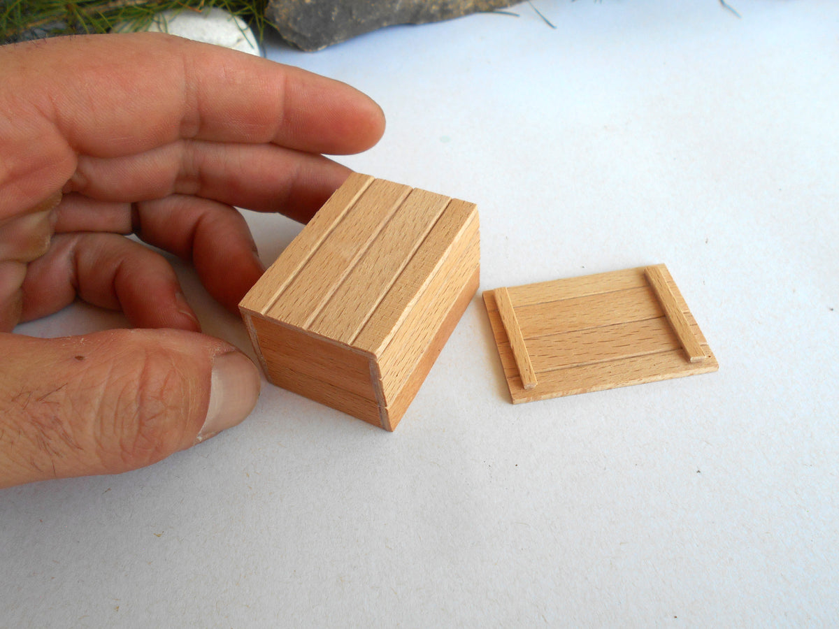 A hand holding a small wooden box next to a matching wooden lid on a light surface.