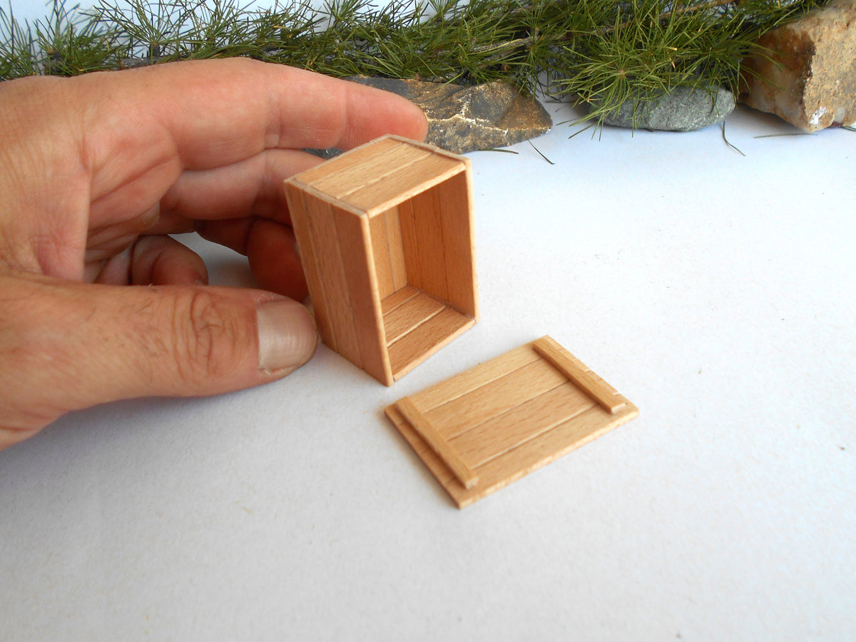 A miniature wooden box with its lid removed, held by a person's hand on a white surface with small rocks and greenery in the background.