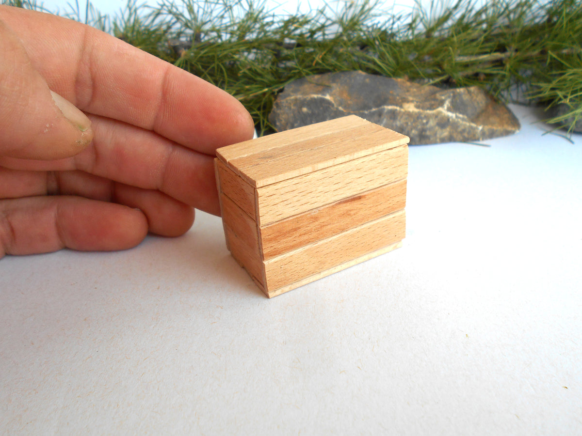A hand holding a small rectangular wooden box with a visible wood grain pattern, set against a white surface with pine branches and a stone in the background.