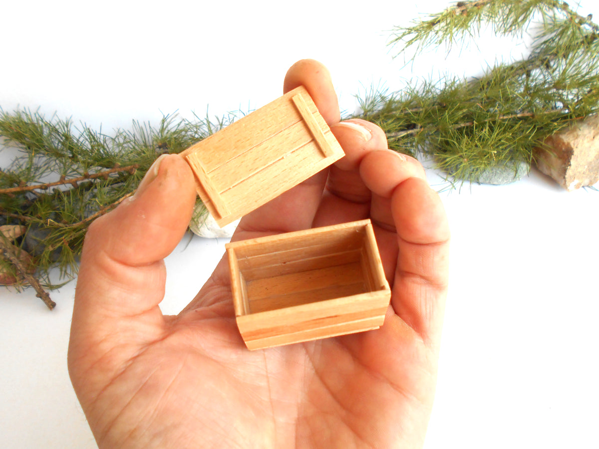 A hand holding a small, open wooden box with a matching lid, shown against a white background with decorative pine branches.