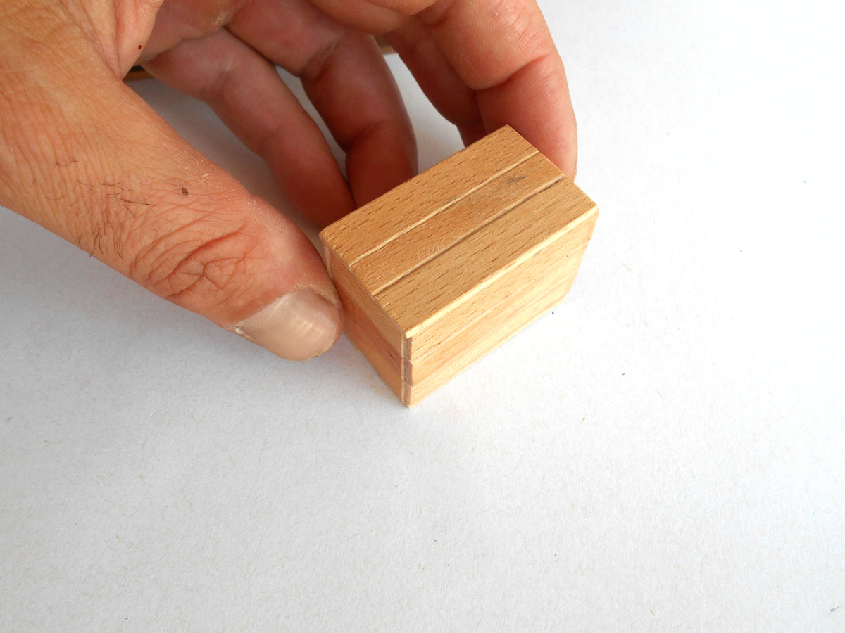 A hand holding a small, light-colored wooden box with a slatted lid against a plain white background.