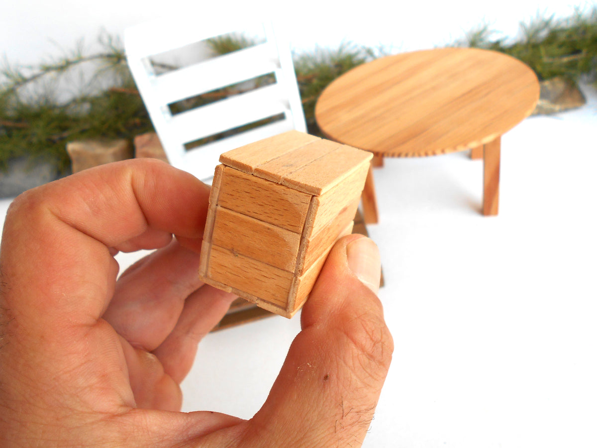 A hand holding a miniature wooden chest or box, with tiny furniture pieces like a round table and chair blurred in the background on a white surface.