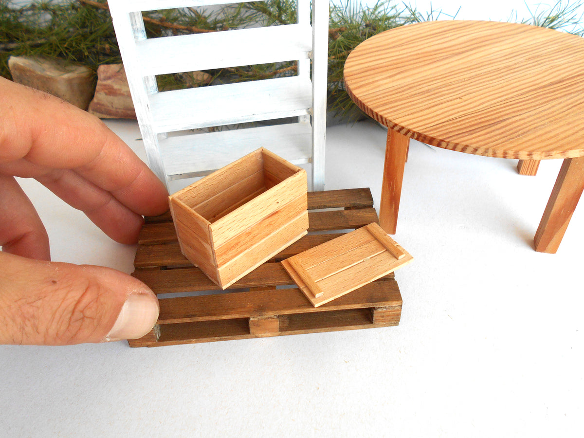 A miniature wooden storage box and matching lid displayed on a tiny wooden pallet, with a human hand holding the pallet for scale. The scene includes a miniature white chair and round wooden table in the background.