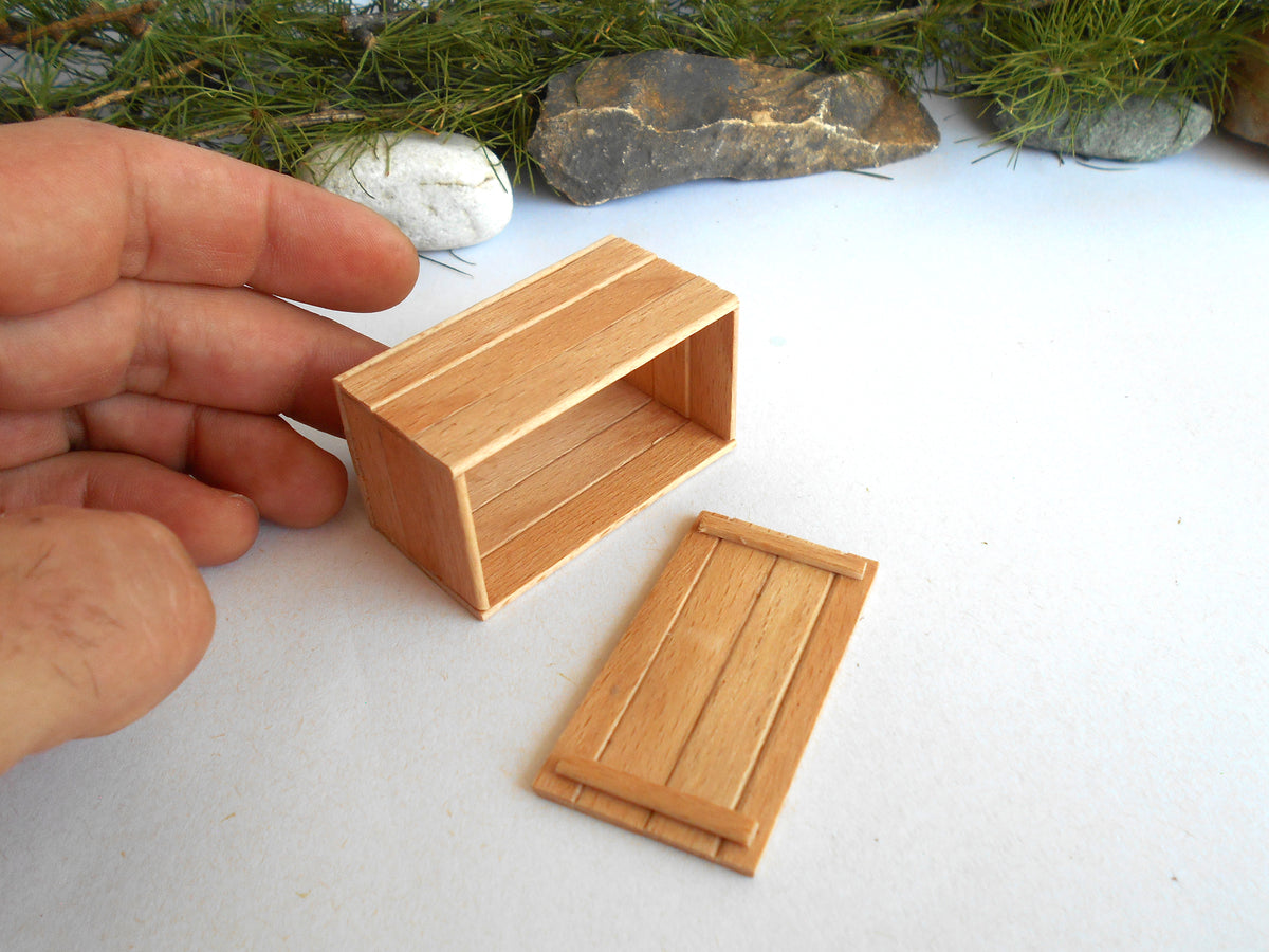A miniature wooden box and lid set, shown with a human hand to indicate scale, on a white surface with decorative stones and pine branches in the background.