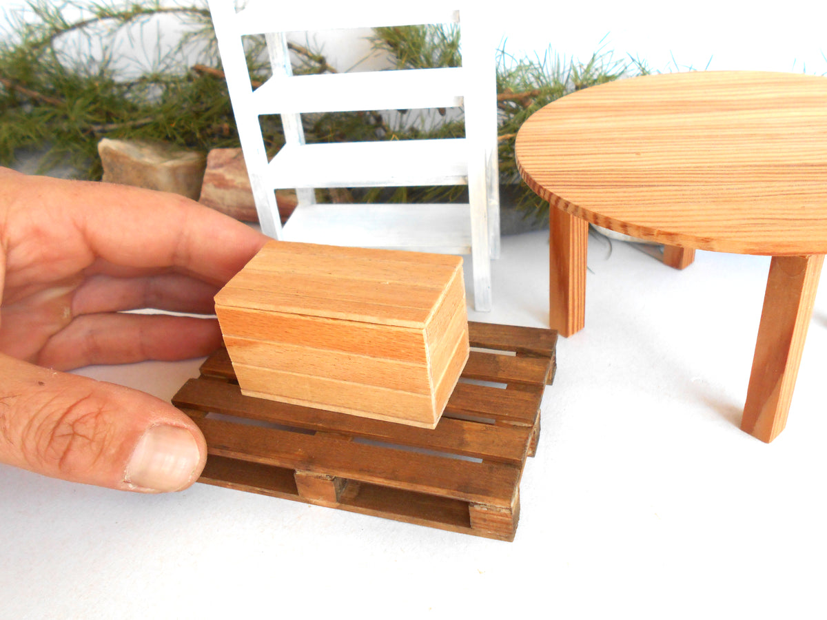 A miniature wooden box resting on a small wooden pallet, with a human hand holding it for scale. The scene also includes a miniature round table and a white chair in the background.