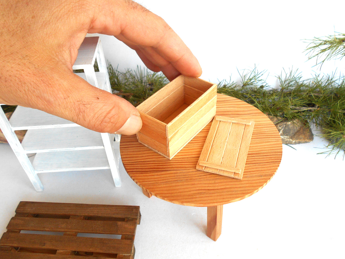 A miniature wooden box set with a lid, shown being held by a human hand to demonstrate scale. The set includes a round wooden table, a small wooden stool, and a wooden pallet, all photographed against a white background with decorative greenery.