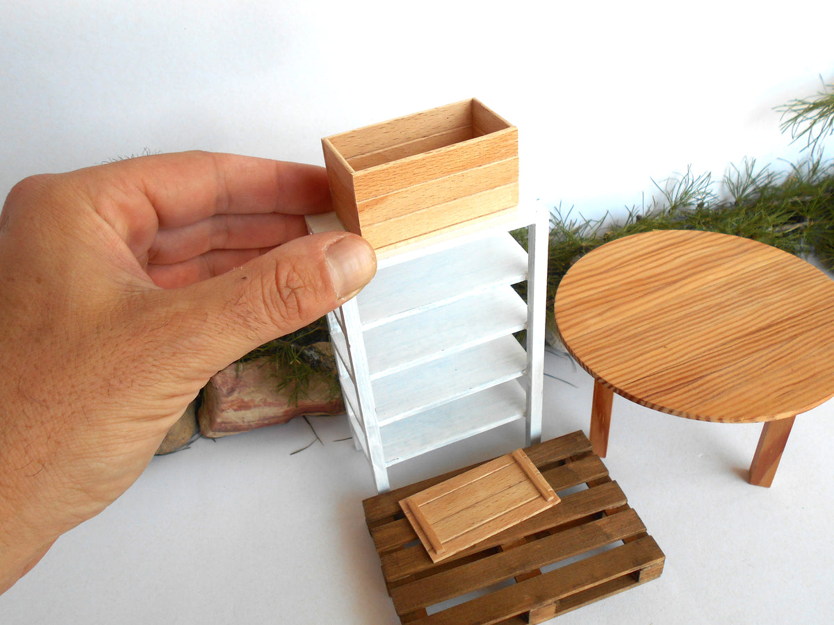 A hand holding a miniature wooden storage box above a small white shelving unit, with additional miniature furniture pieces including a round wooden table and a slatted wooden crate on a white surface.