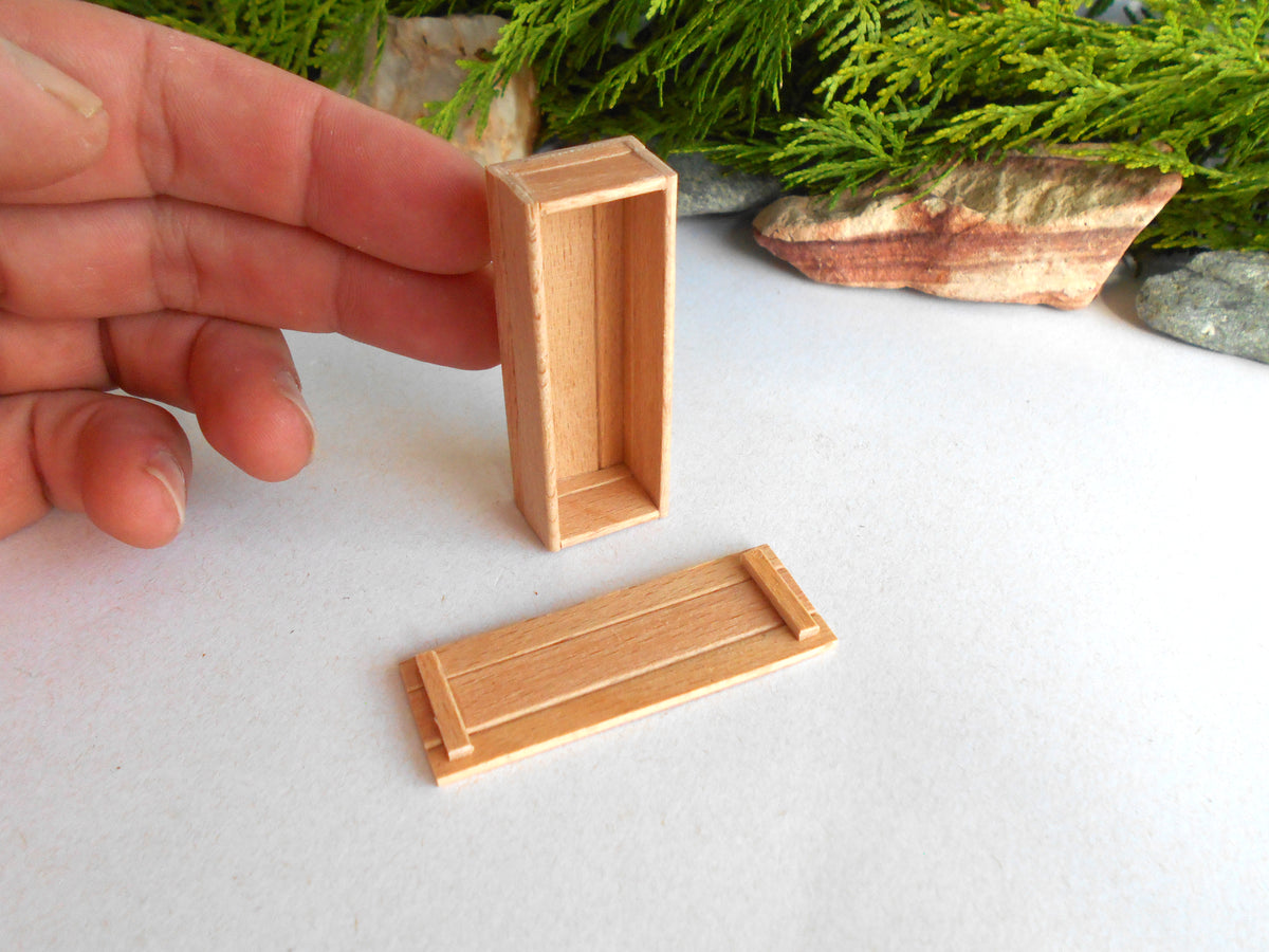 A hand holds a small rectangular wooden box with a separate matching lid or cover placed below it on a white surface, with green foliage and stones in the background.