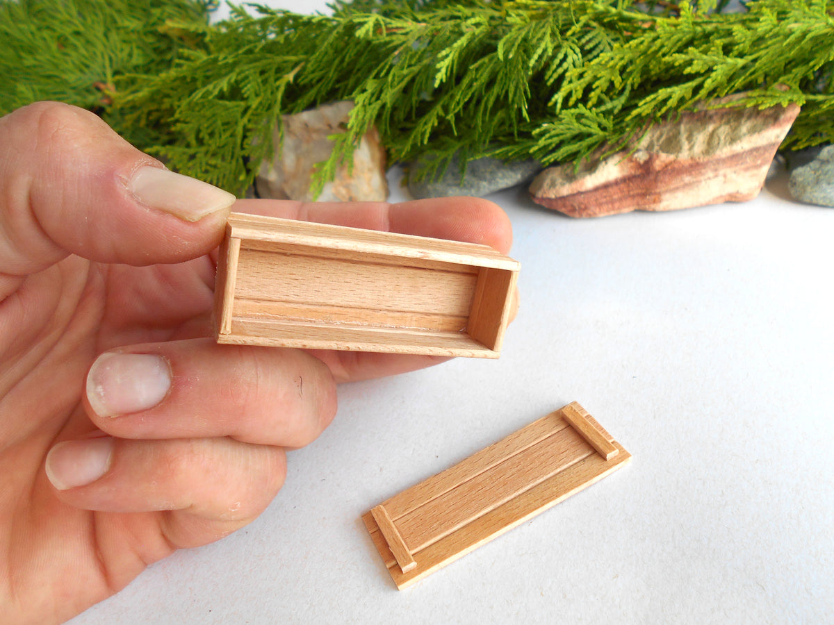 A hand holds a small rectangular wooden box with a hinged lid, while another matching wooden box lies on a white surface below. Green foliage and stones are blurred in the background.
