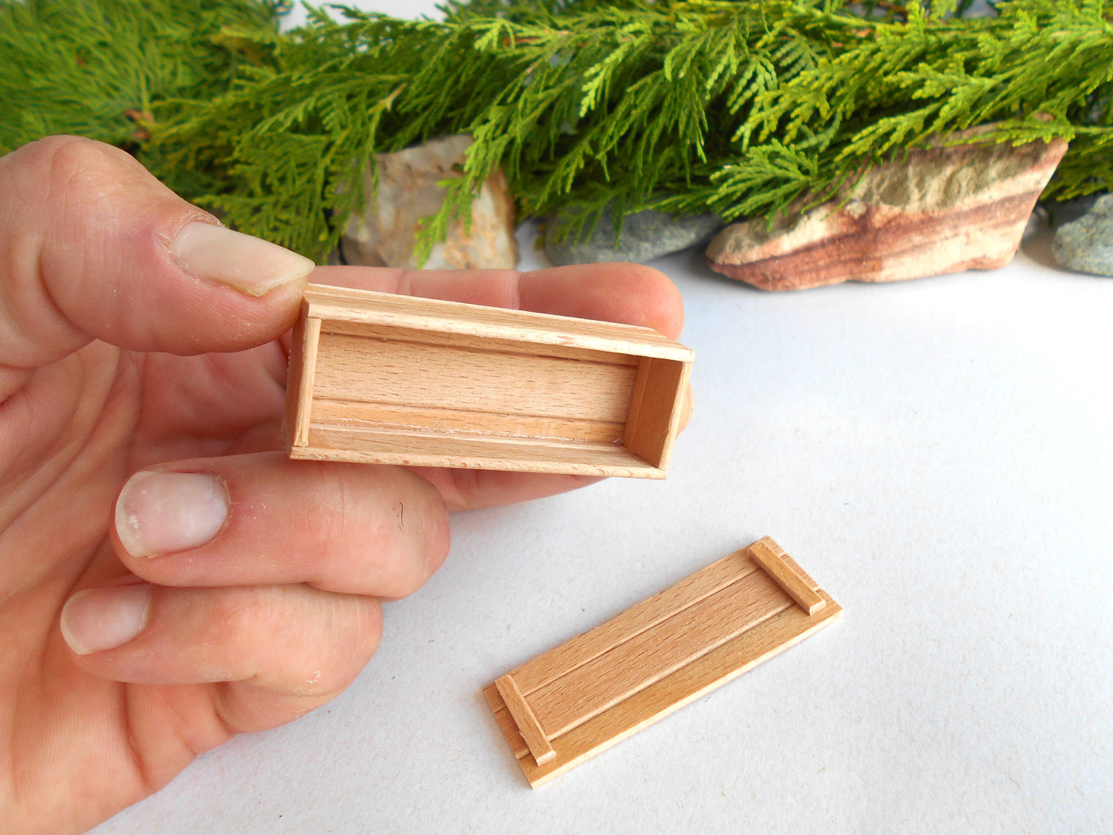 A hand holds a small rectangular wooden box with a hinged lid, while another matching wooden box lies on a white surface below. Green foliage and stones are blurred in the background.