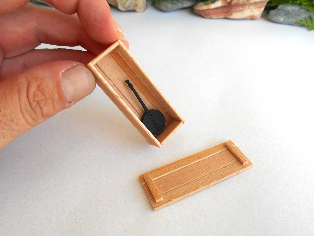 A hand holding a miniature wooden box with a small black object inside, with the matching wooden lid placed below on a white surface.