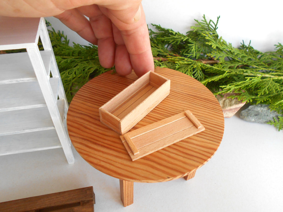 A close-up of a miniature wooden table with a round wood-grain top and small legs, holding two small rectangular wooden boxes. A human hand is visible holding one of the boxes, and green foliage decor is in the background.