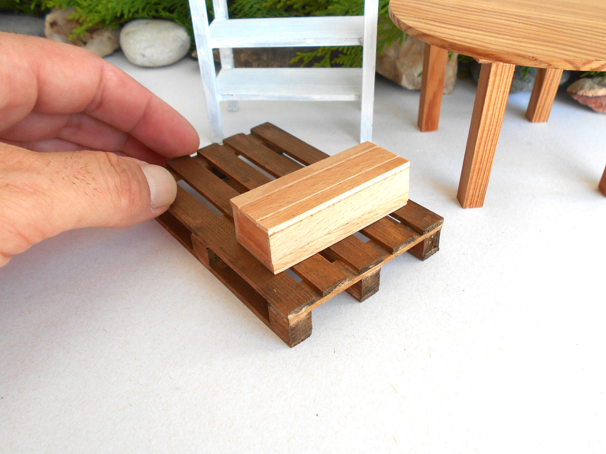A miniature wooden pallet and matching block, likely for dollhouse or scale model furniture, with a human hand adjusting it on a white surface.