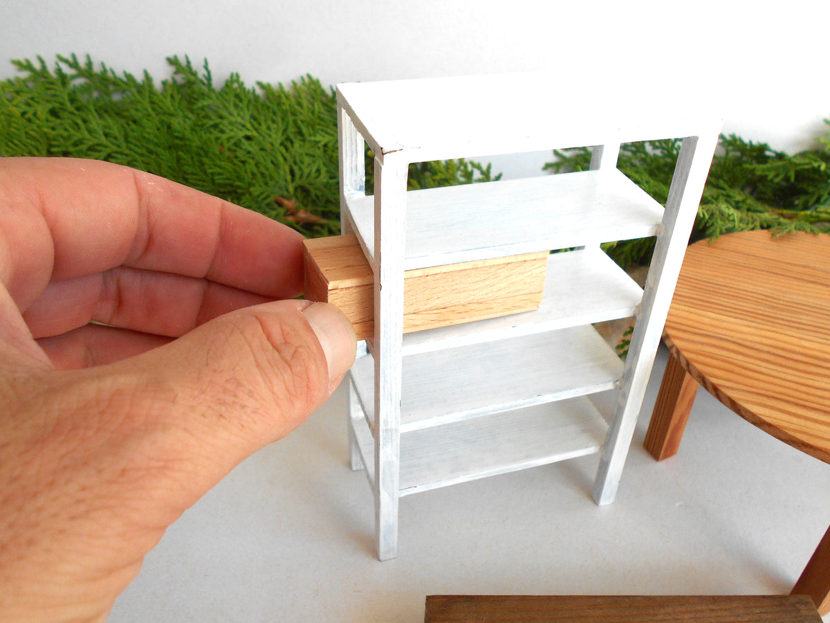 A hand holding a small wooden block next to a miniature white shelving unit with a matching wood drawer, set against a backdrop of green foliage and a wooden table.