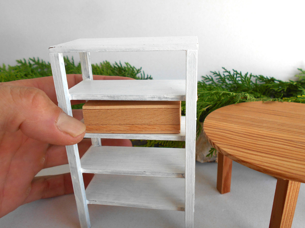 A miniature white shelving unit with a small wooden drawer, held by a hand to show scale, with a miniature round wooden table and green plants in the background.