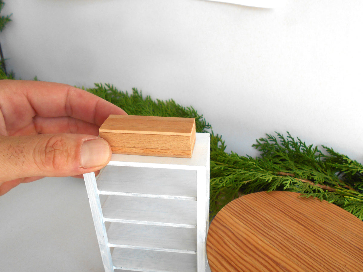 A hand holding a small rectangular wooden box above a miniature white shelving unit, with green foliage and a round wooden table in the background.