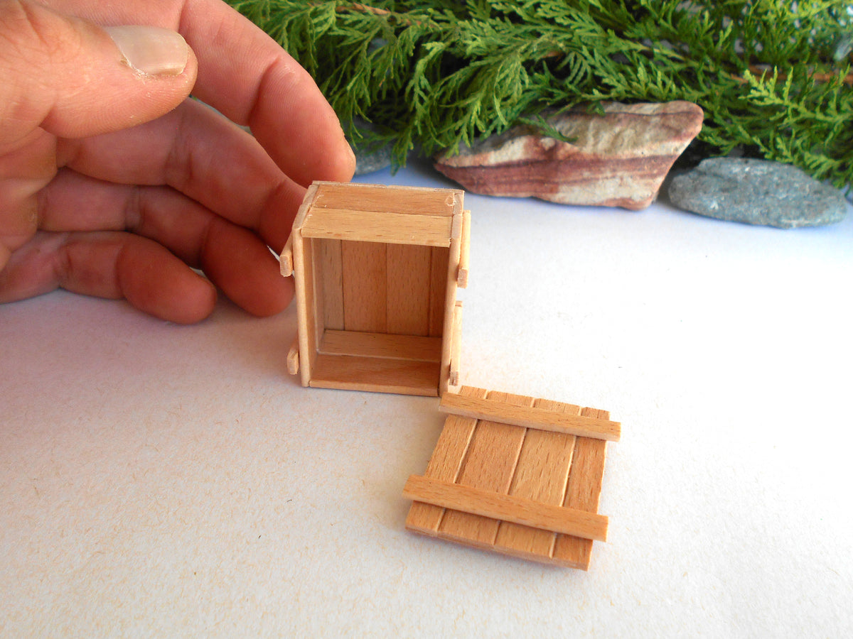 A miniature wooden crate or box with its lid placed beside it, shown on a white surface with a hand holding one side. Greenery and stones are blurred in the background.