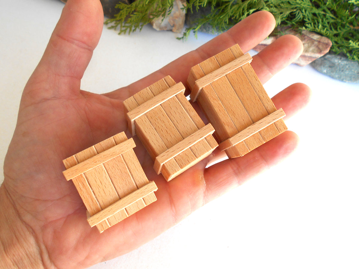 Three miniature wooden crates in different sizes held in a person's hand, shown against a white background with some greenery and rocks blurred in the upper background.