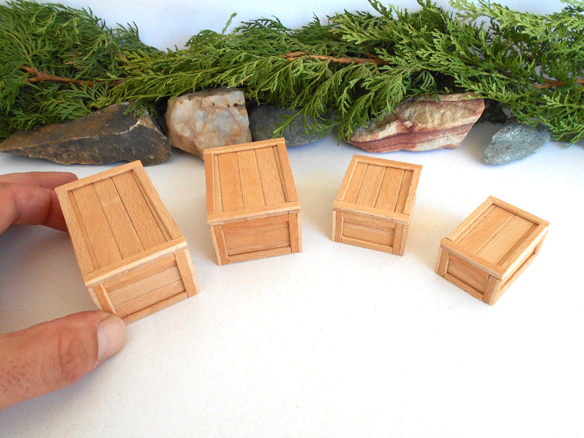 Four miniature light wood crates of different sizes arranged on a white surface, with a hand holding one crate on the left and green foliage and rocks in the background.