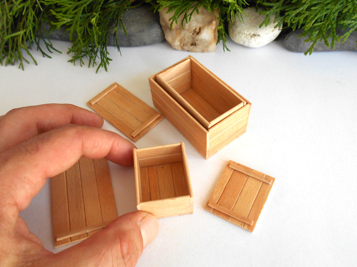 A hand holding a miniature wooden box with a matching lid, surrounded by other small wooden boxes and lids on a white surface with green foliage and stones in the background.