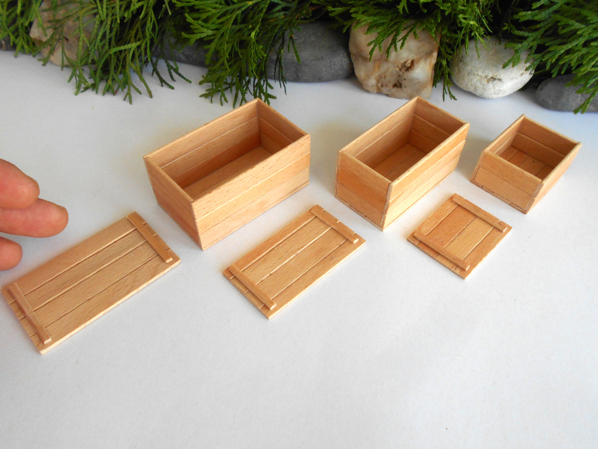 Three miniature light wood boxes and lids arranged on a white surface, with a hand partially visible on the left and greenery in the background.