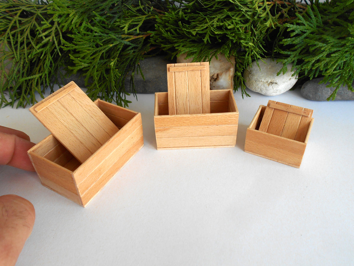 Three miniature light wood boxes of different sizes arranged on a white surface with green foliage in the background. A human hand is visible holding the leftmost box open.