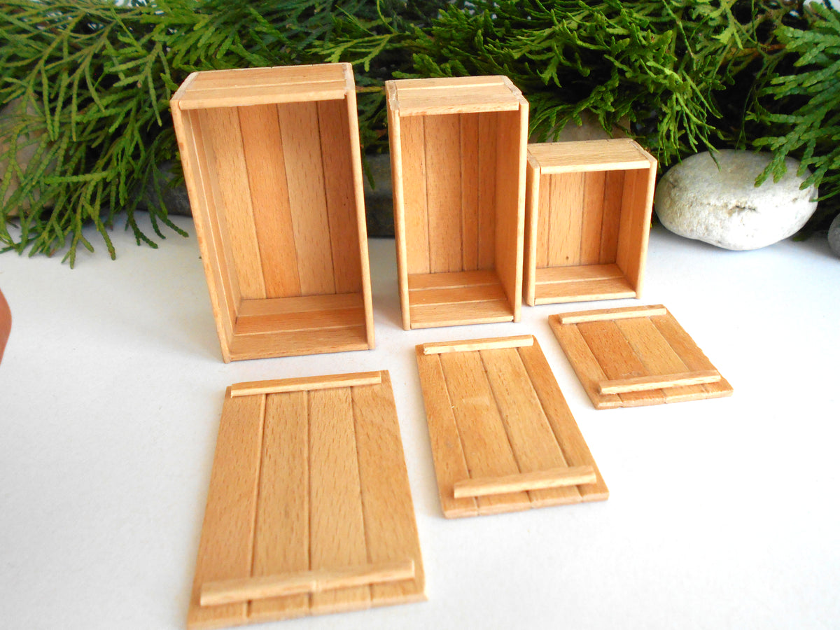 A set of miniature wooden storage boxes and lids, likely for dollhouse or model building, made from light natural wood. The set includes three tall rectangular boxes, two square boxes, and four slatted lids, arranged on a white surface with green foliage in the background.
