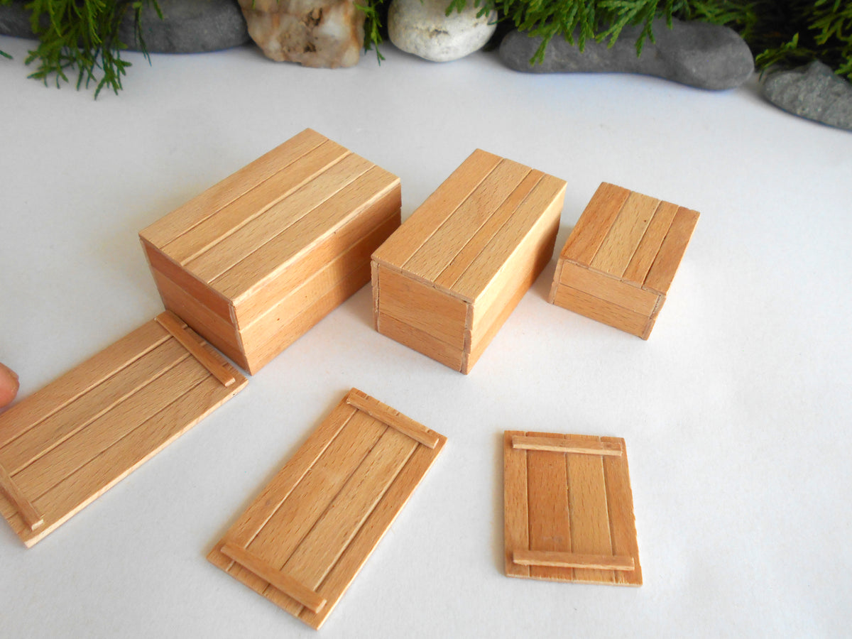 Three miniature light wood boxes and lids arranged on a white surface, with a hand partially visible on the left and greenery in the background.