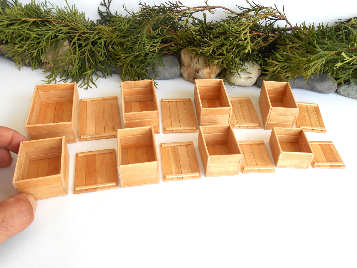 A set of miniature light wood crates and boxes arranged on a white surface, with a hand holding one box at the left edge and evergreen branches and stones in the background.