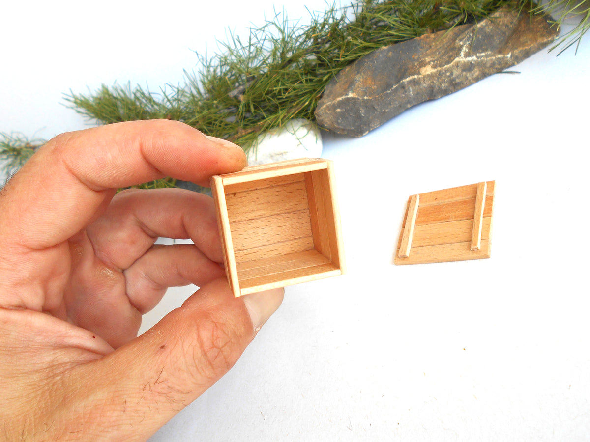 A hand holding a small square wooden box with its lid placed beside it on a white surface, with greenery and a stone in the background.