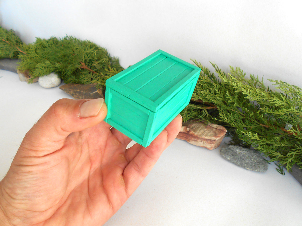 A hand holding a small, bright turquoise-green miniature wooden chest or box, set against a white tabletop with decorative green moss and small stones.