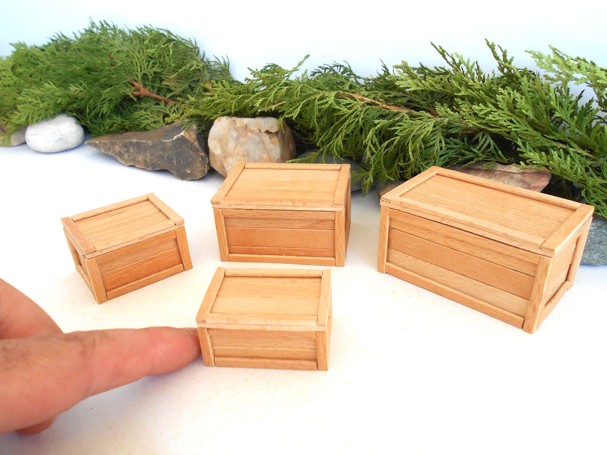 Four miniature light wood crates of different sizes displayed on a white surface with green pine branches and small rocks in the background. A human finger is visible in the bottom-left corner for scale.