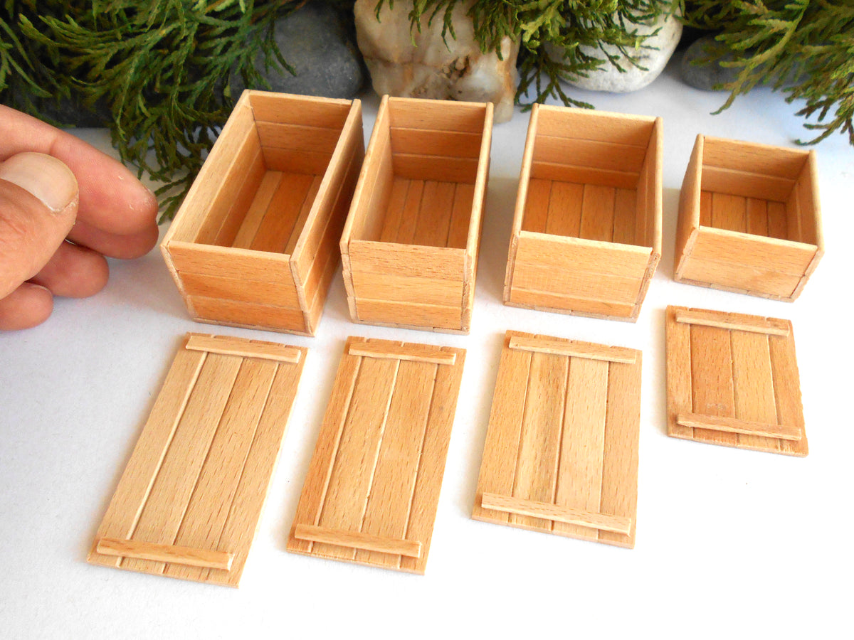 A set of miniature light wood crates and lids, likely for scale models, arranged on a white surface with a hand holding one crate on the left.