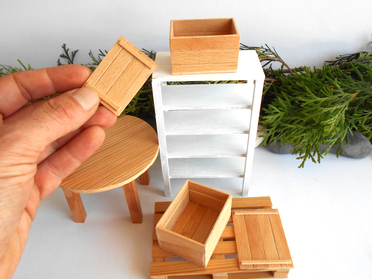 A hand holds a small wooden crate in front of a miniature white shelf and other dollhouse-style wooden furniture pieces, including a round stool and another crate on a pallet.