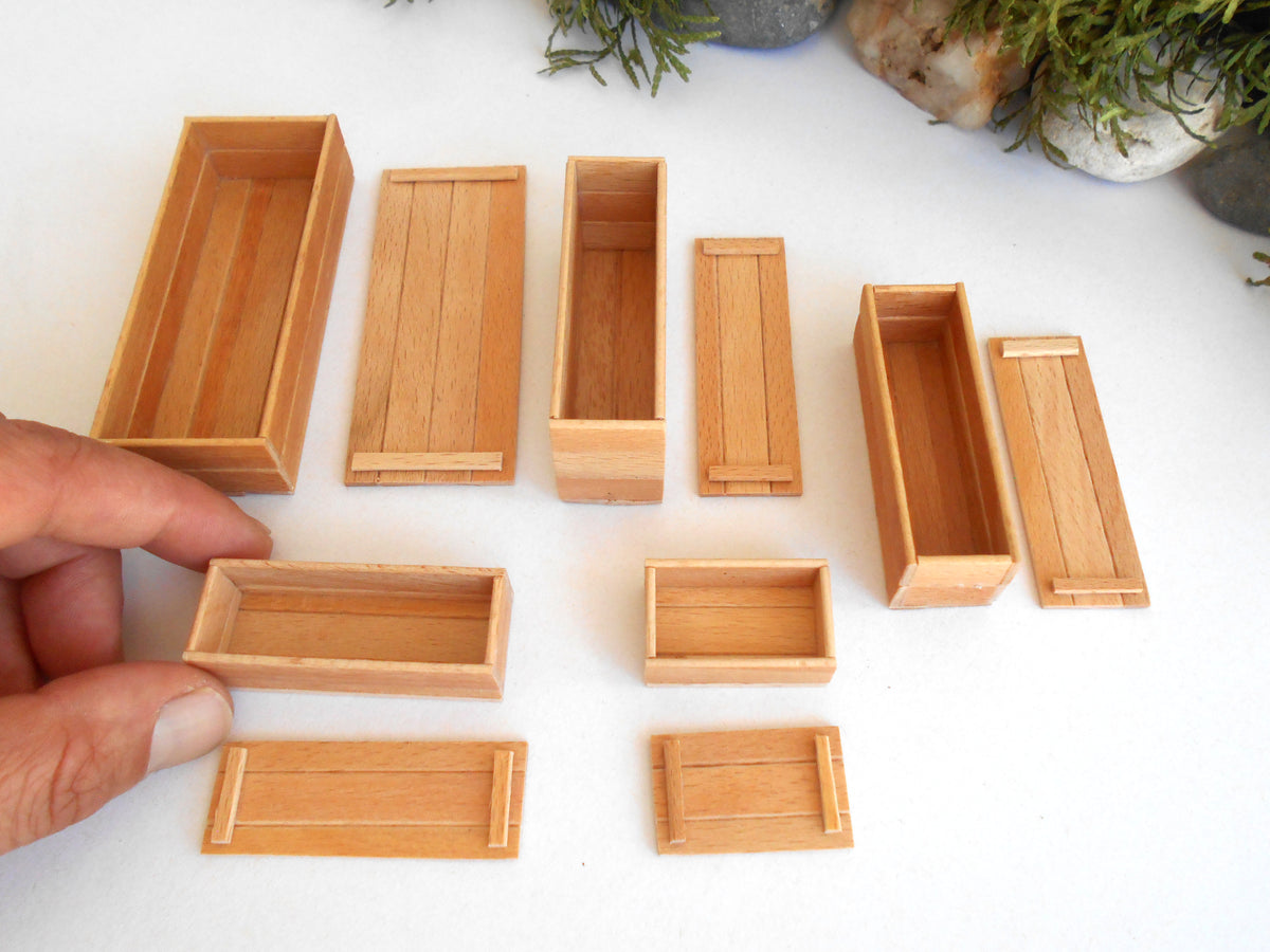 A set of miniature rectangular wooden boxes in various sizes, shown on a white surface with a hand holding one of the smaller boxes. Small plants are visible in the background.