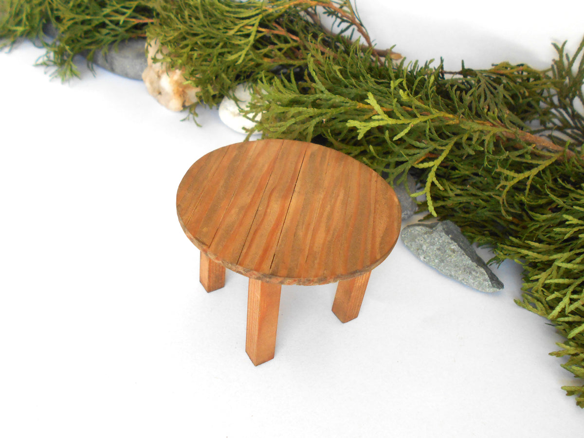 A miniature round wooden stool or table with four legs, styled with green foliage and small stones on a white background.