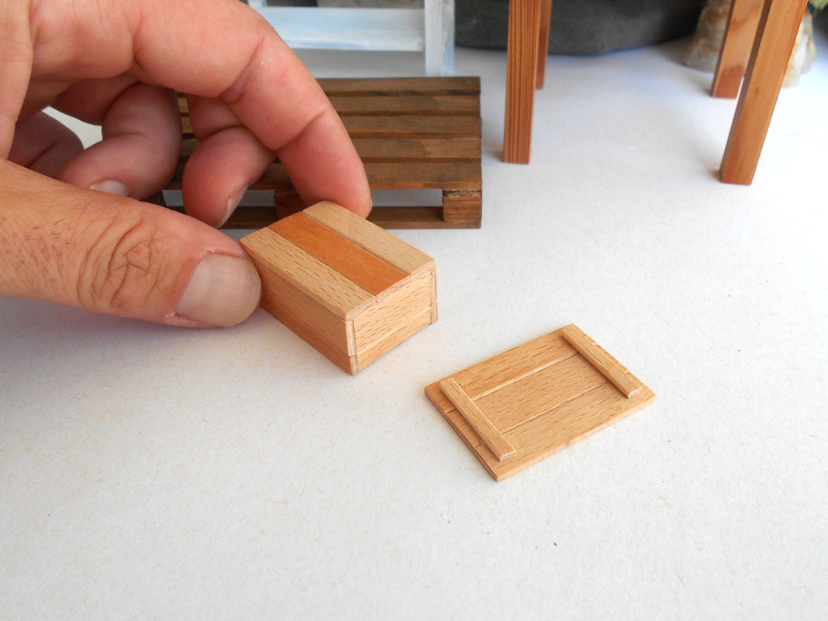 A close-up of a hand holding a small wooden miniature box with a striped pattern, next to a matching flat wooden piece on a white surface.
