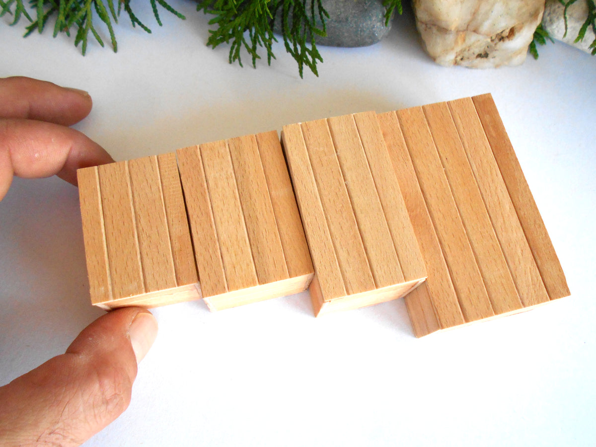A hand holding a set of rectangular wooden blocks with grooved sides, displayed on a white surface with decorative greenery in the background.