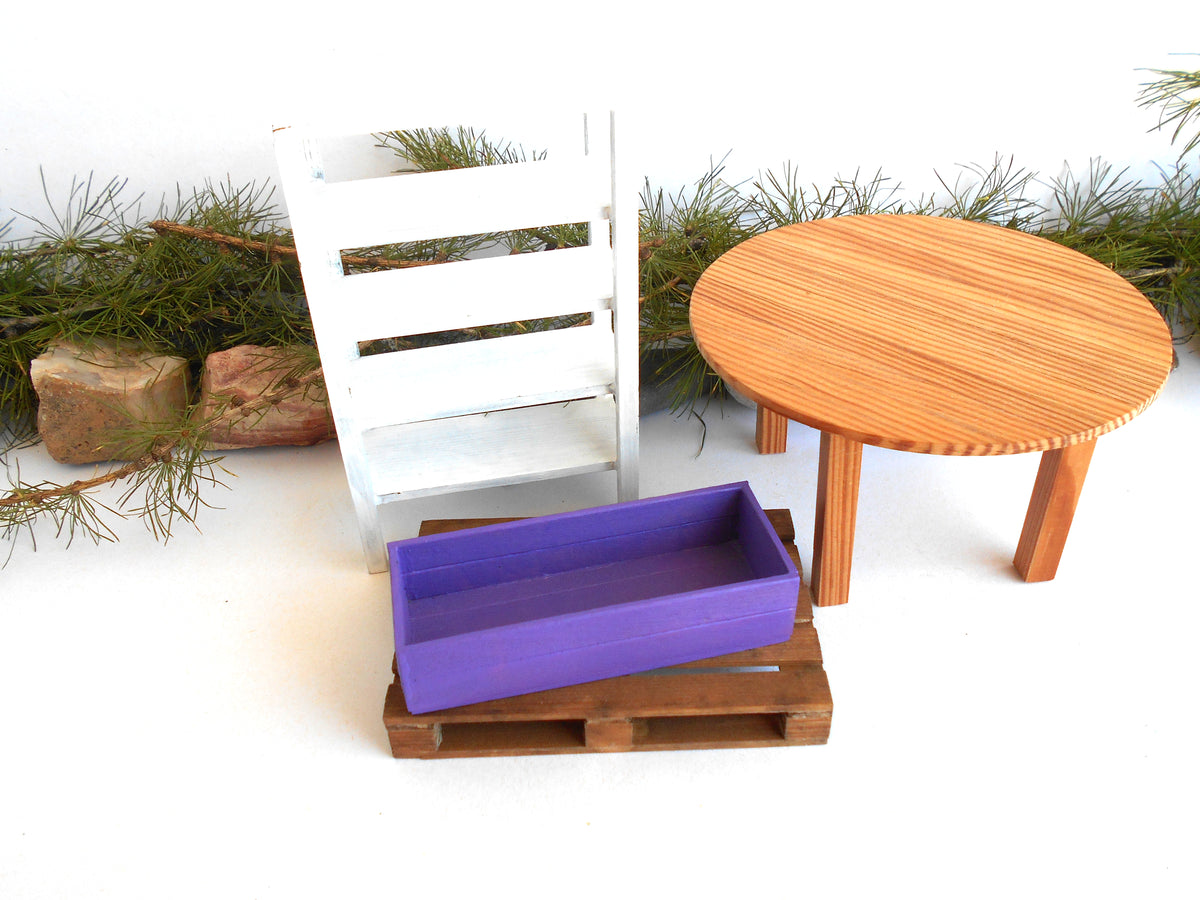 A miniature dollhouse furniture set featuring a purple rectangular tray on a brown wooden pallet base, a white slatted chair, and a round light brown wooden table, styled with pine branches and small rocks on a white background.