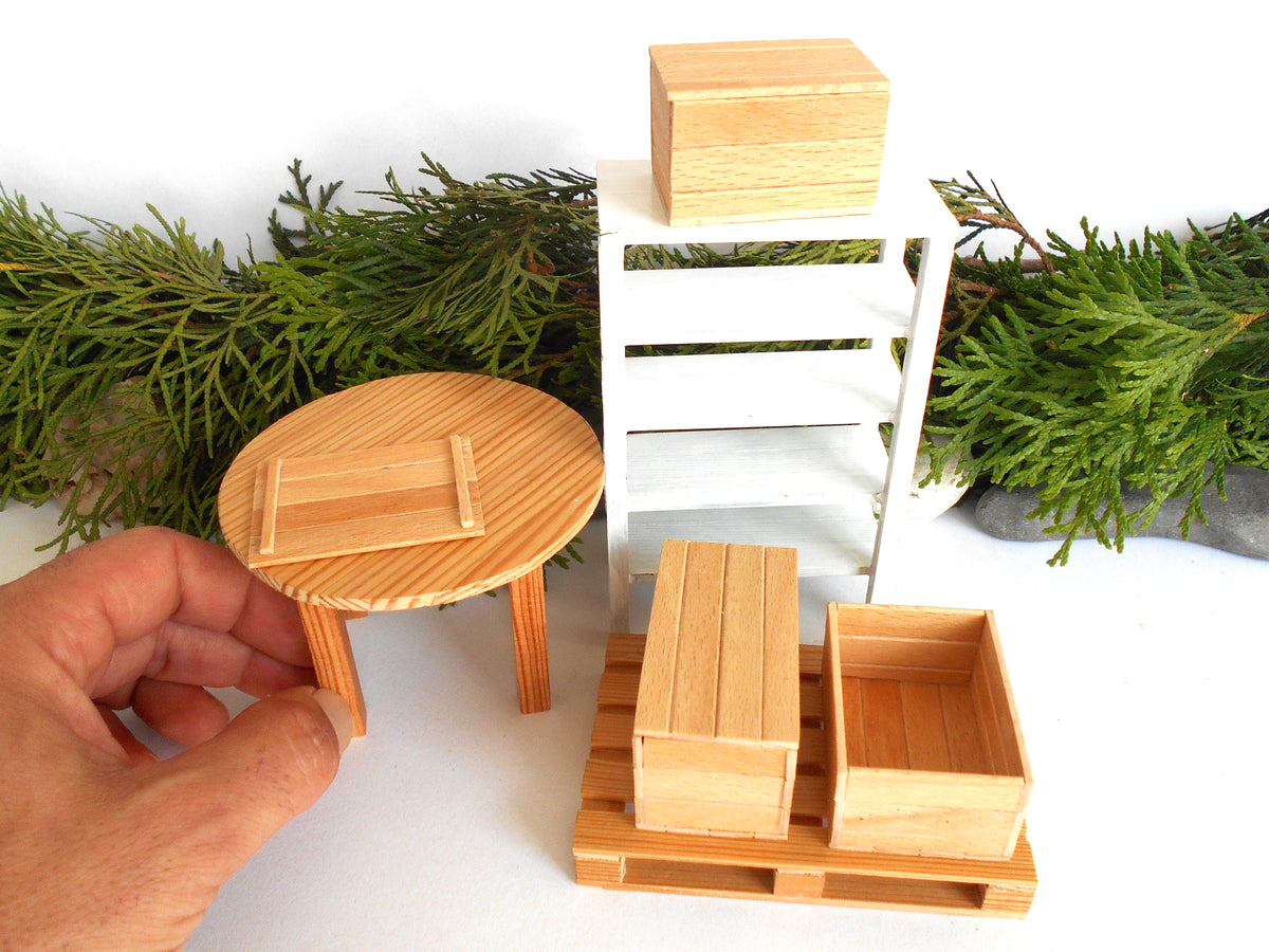 A miniature wooden dollhouse furniture set including a round table held by a hand, a white shelving unit, and two storage crates on a pallet, displayed with green pine branches against a white background.