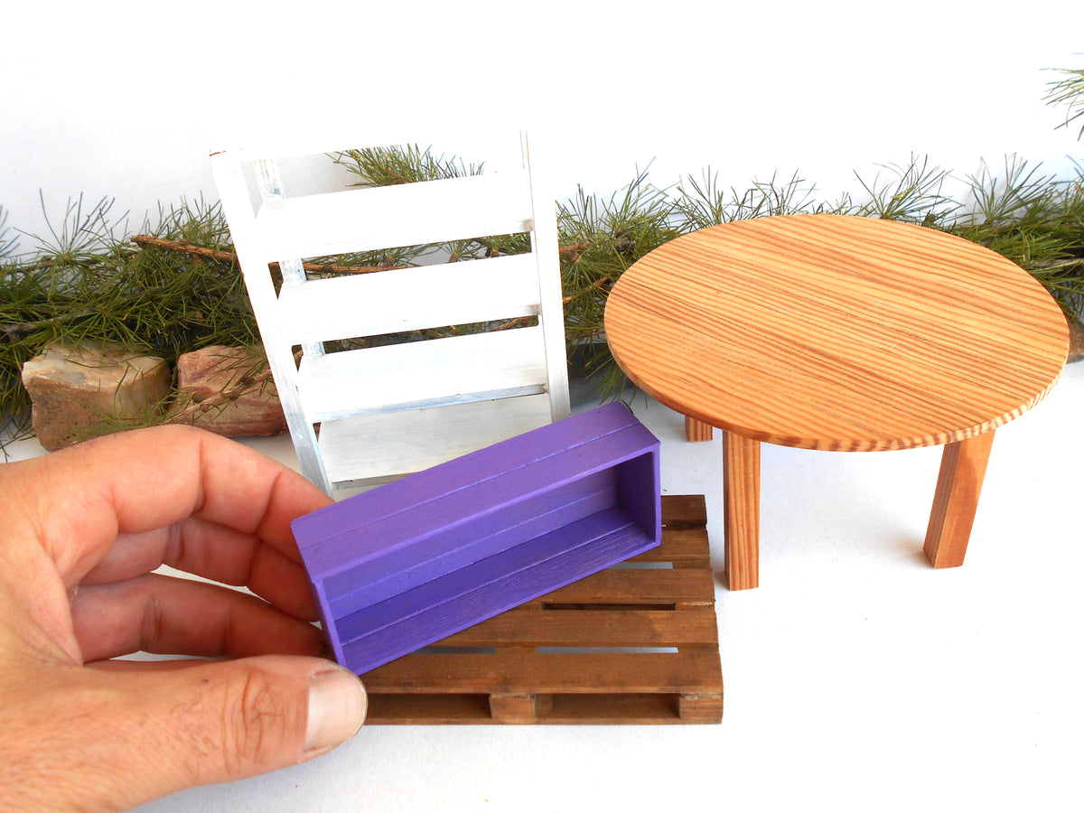 A hand holds a miniature purple storage box in front of a miniature wooden table and chair set, likely for dollhouse or scale model use.