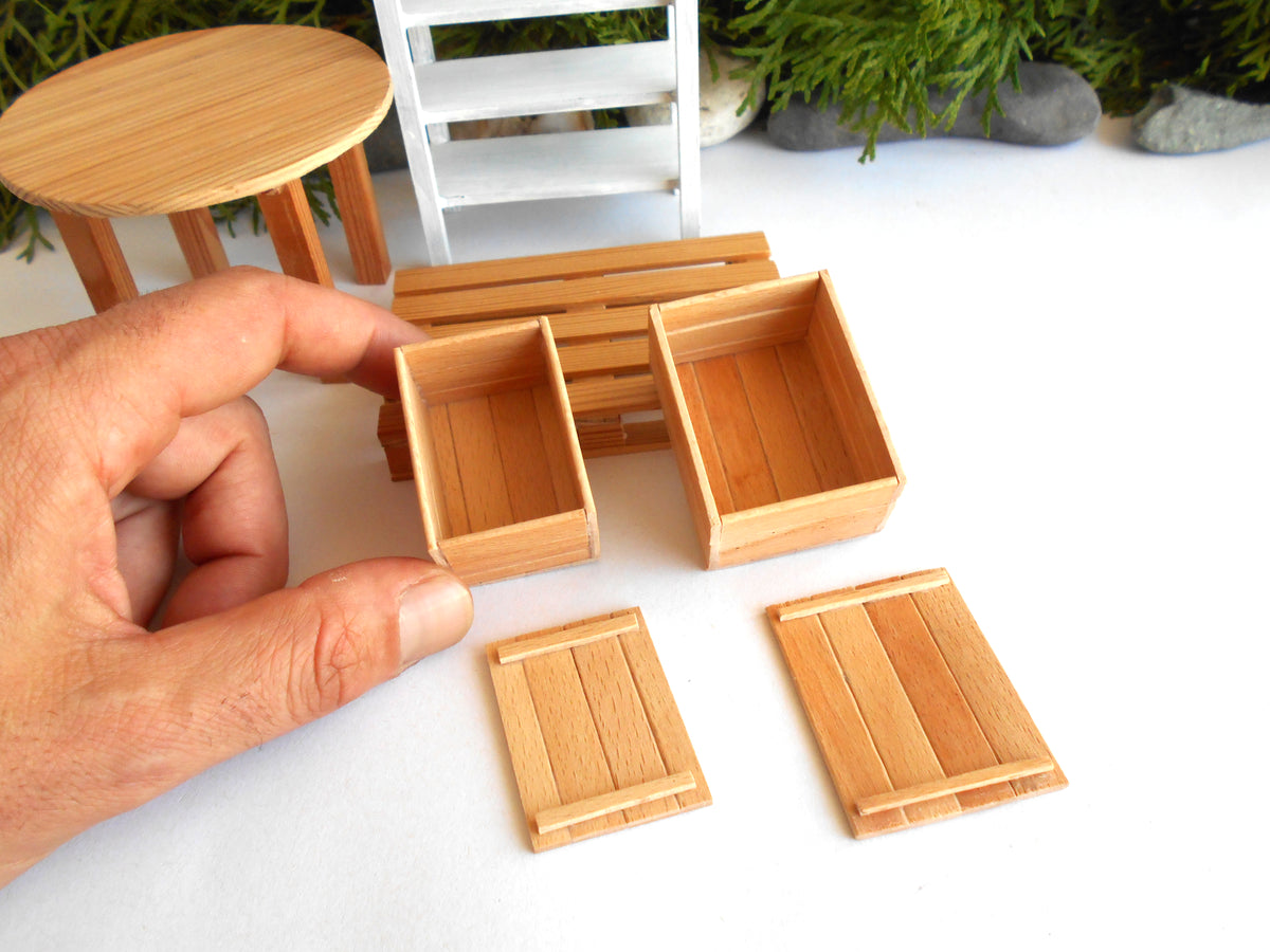 A hand holding a miniature wooden crate box, with several other small wooden crate and bench pieces arranged on a white surface. The background shows a blurred miniature white chair and greenery.