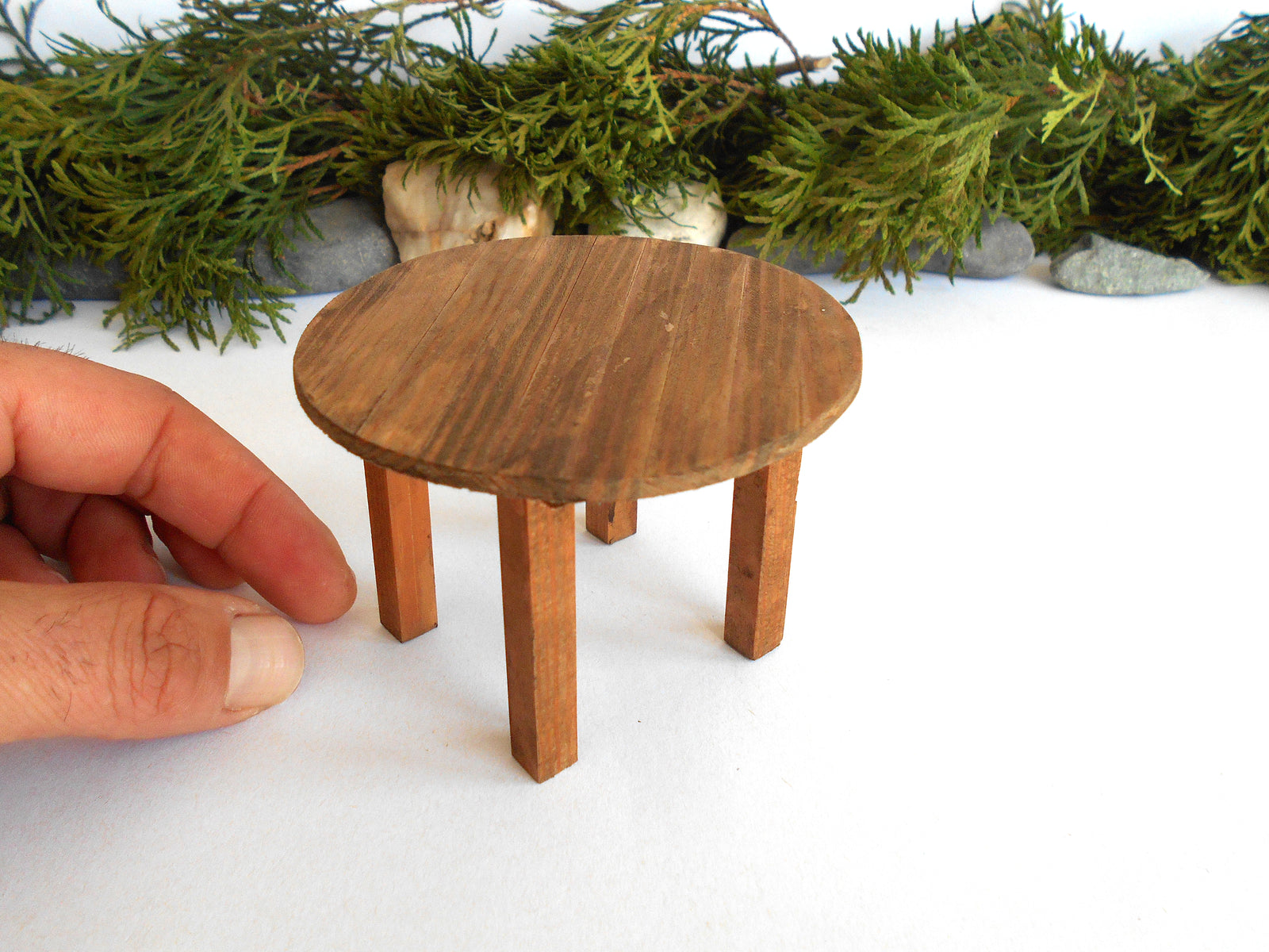 A miniature round wooden table with four legs, shown next to a human hand to demonstrate its small scale, set against a white background with green foliage at the top.