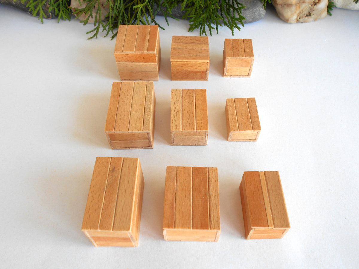 Nine small rectangular wooden boxes in varying sizes, arranged in three rows on a white surface with green foliage and stones in the background.