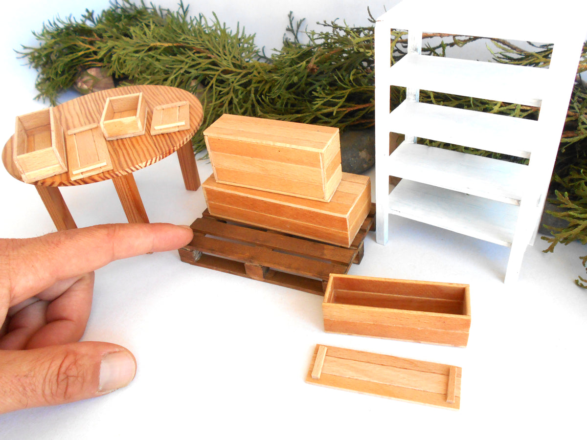 A miniature wooden toy furniture set including a small oval table with three cubicles, a stack of wooden boxes on a pallet, a white shelving unit, and two long rectangular boxes, shown with a person's hand for scale against a white background with greenery.
